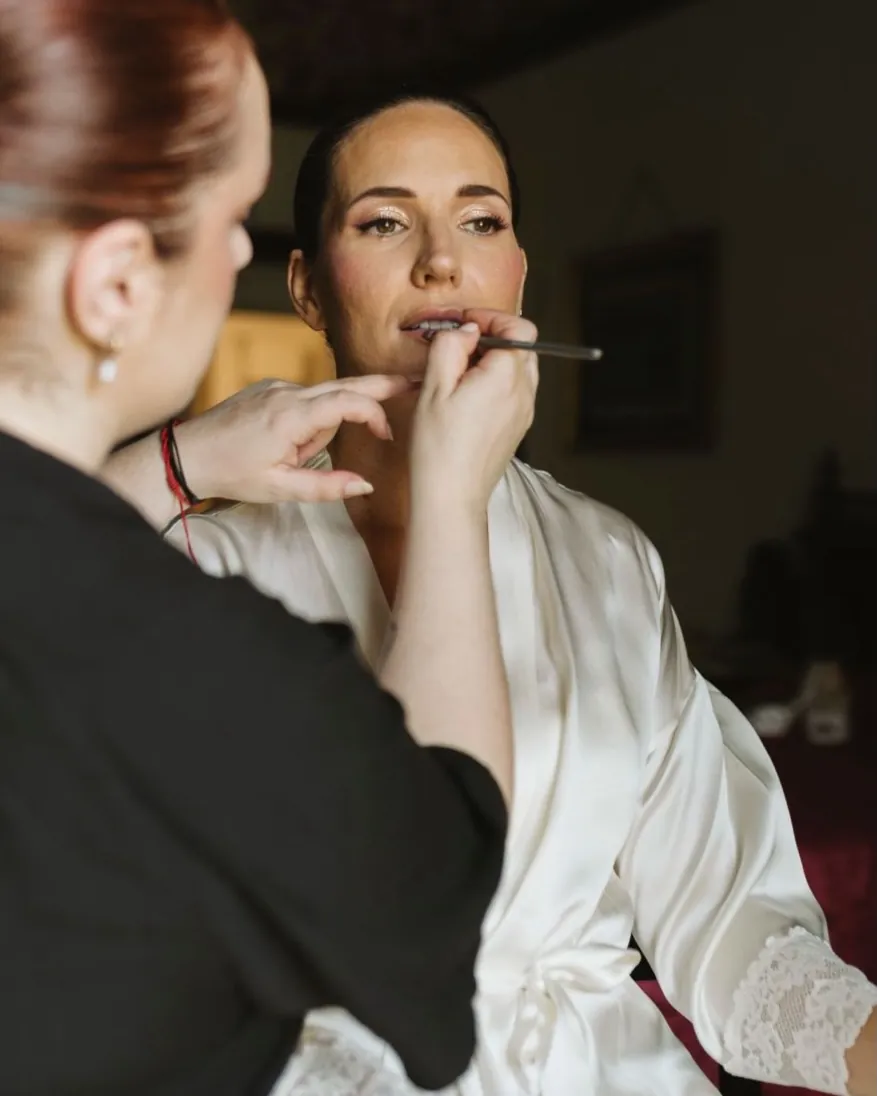 Ester maquillando los labios a una novia en bata de satén blanca, junto a una ventana