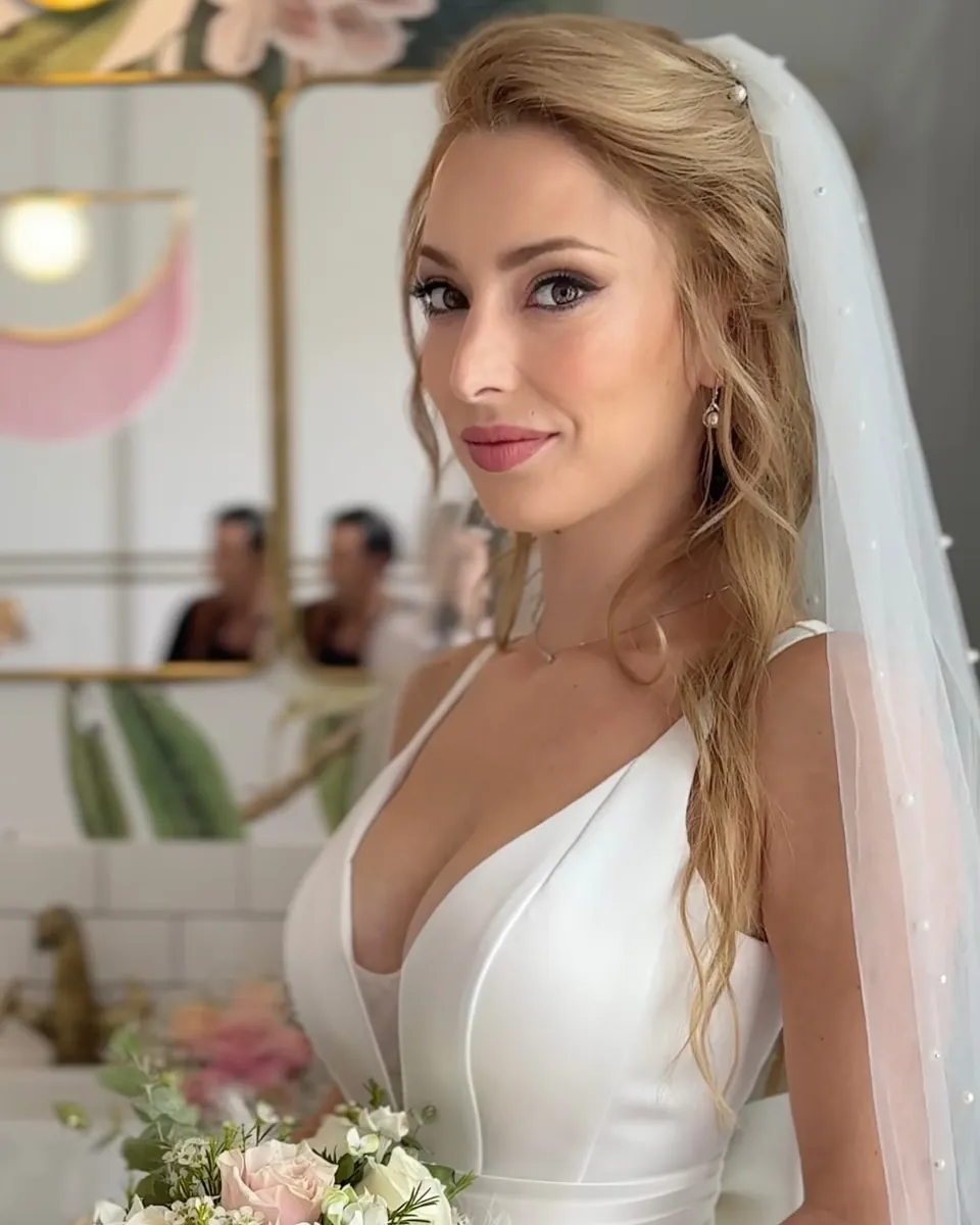 Novia rubia con velo largo y ramo de rosas, de perfil mirando a cámara